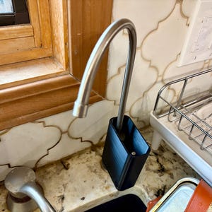 May include: A stainless steel faucet with a black rectangular holder. The faucet is curved and positioned over a sink. The holder is placed on a countertop with a decorative tile backsplash.