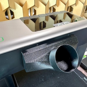 May include: A silver and black 3D printer with a black cylindrical nozzle. The printer has a rectangular green light and several buttons on top. The nozzle is attached to a black rectangular component.