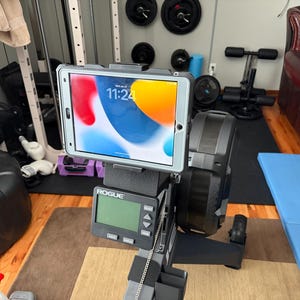 May include: A gray tablet secured to a Rogue rowing machine, displaying a colorful screen with the time 11:24. The machine has a black display panel and is set up in a home gym setting with weights and exercise equipment in the background.
