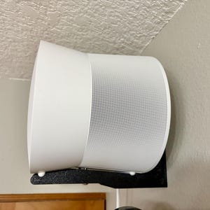 May include: A white cylindrical speaker mounted on a black metal bracket. The speaker has a perforated front panel and is positioned in a corner against a light-colored wall. The bracket is attached to the wall.