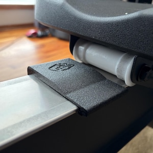 May include: Close-up of a rowing machine component. A textured, dark gray metal piece with a hand symbol is attached to a silver rail. A white cylindrical component is visible, along with a black seat.