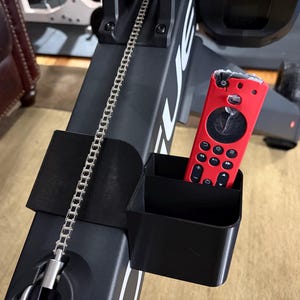 May include: A black exercise machine with a silver chain and a red remote control in a black holder. The remote has multiple black buttons and a circular control. The machine has white lettering.