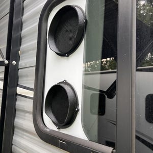 May include: A white rectangular panel installed within a recreational vehicle window frame, featuring two circular black air vents with mesh screens. The vents are stacked vertically, and the entire assembly is set against a gray corrugated metal wall.