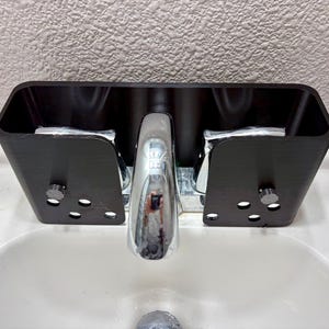 May include: A black, rectangular sink organizer with a curved top and sides, designed to fit around a faucet. The organizer has several small, oval-shaped holes and is mounted on a white sink. The faucet is chrome.