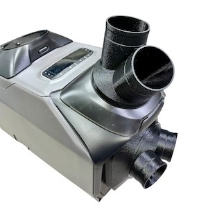 May include: A gray and black ventilation system with multiple black cylindrical outlets. The device has a rectangular shape with a curved top and a control panel. The outlets are positioned at different angles, suggesting airflow direction.