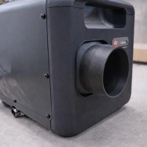 May include: A dark gray industrial machine with a handle and a large cylindrical outlet. The machine has a boxy shape with rounded corners and visible screws. The outlet is a dark gray color.