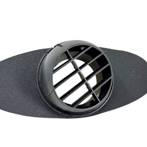 May include: A black plastic air vent with a circular, slotted design. The vent is mounted on a textured, oval-shaped black base. The design features a series of horizontal and vertical bars within the circular opening.
