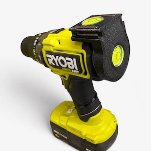 May include: A lime green and black Ryobi One HP cordless drill with a black 3D-printed attachment mounted on the back. The attachment features two integrated circular bubble levels, one positioned on the top and one on the rear face.