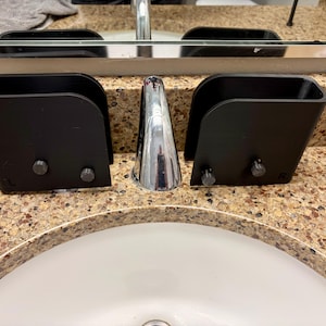 May include: Two black, wall-mounted toothbrush holders with a curved design. Each holder is secured with two silver bolts. The holders are positioned on a speckled countertop near a chrome faucet and a white sink.