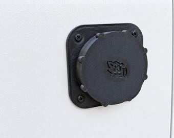 May include: A black, circular, 3D-printed plastic cap with a textured surface and a small embossed graphic in the center. The cap is mounted to a square base plate with rounded corners, secured by four black screws against a white surface.