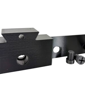 May include: Black 3D-printed mechanical component with a rectangular base, a notched top, and a cylindrical hole. A flat, rectangular piece extends from the base. Two screws and a cylindrical peg are also present. The components are designed for mechanical use.