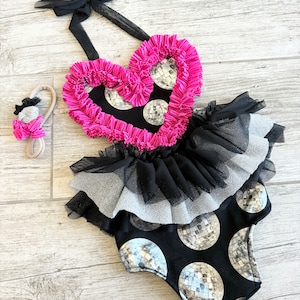 May include: A black and pink baby onesie with a heart-shaped top and a silver disco ball print. The onesie has a black tulle skirt and a matching headband with a silver disco ball.