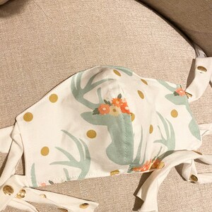 May include: White face mask with a green deer print and gold polka dots. The mask has white ties.