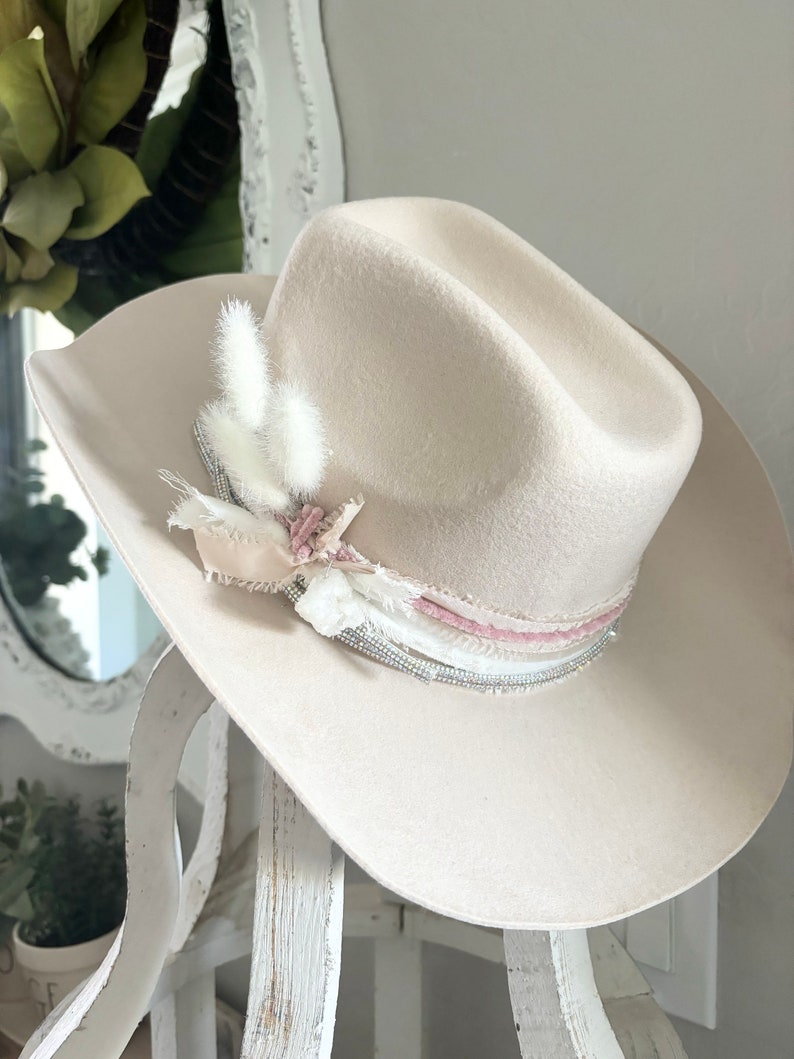 May include: A cream-colored felt cowboy hat with a wide brim. The hat is decorated with a pink and white ribbon, white feathers, and a silver beaded trim.