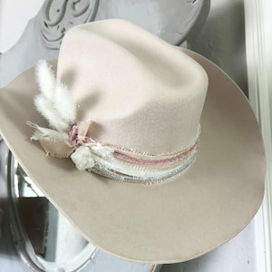 May include: A light beige cowboy hat with a wide brim and a pink and white feather trim. The hat has a beaded band and a pink ribbon.