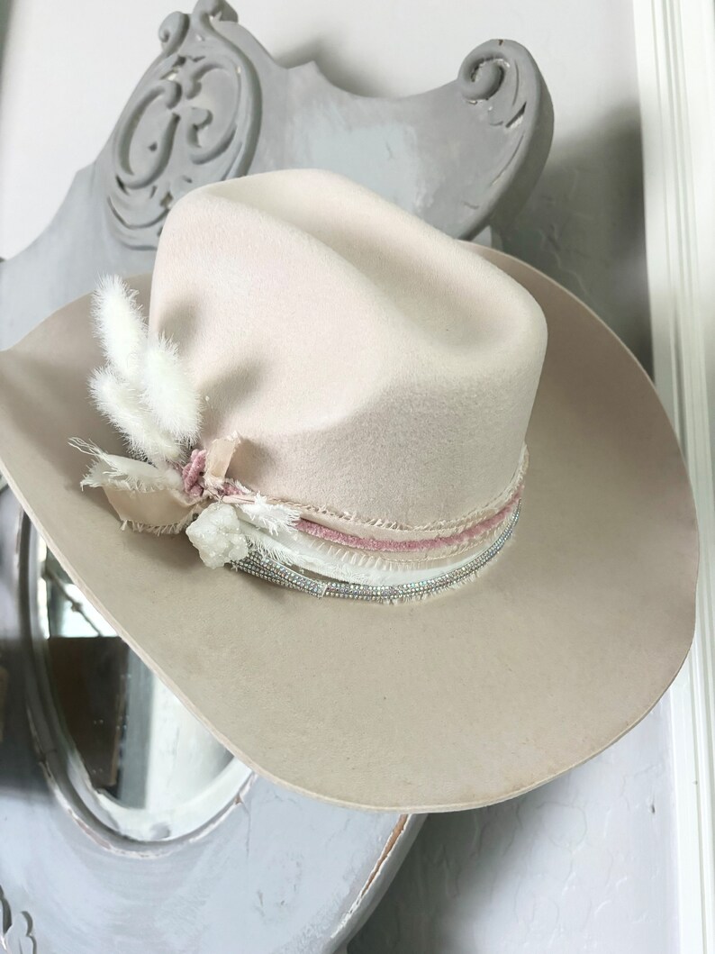 May include: A light beige cowboy hat with a wide brim and a pink and white feather trim. The hat has a beaded band and a pink ribbon.