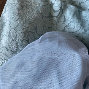 May include: A light blue tulle fabric with a fine mesh pattern. The fabric is draped over a light blue patterned fabric with a floral design.