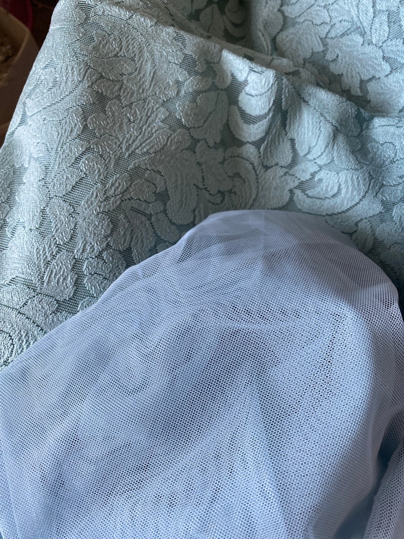 May include: A light blue tulle fabric with a fine mesh pattern. The fabric is draped over a light blue patterned fabric with a floral design.