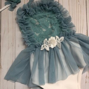 May include: A baby girl's outfit with a white bodysuit and a blue tulle skirt with lace overlay. The outfit includes a matching headband with a pearl accent.