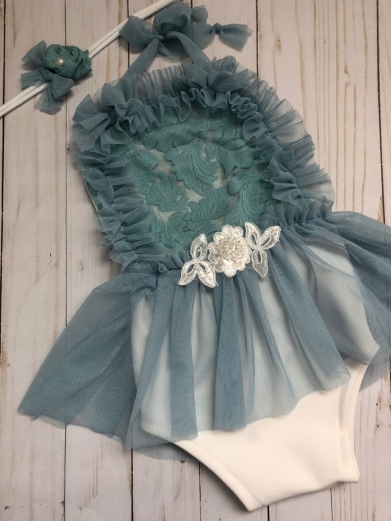 May include: A baby girl's outfit with a white bodysuit and a blue tulle skirt with lace overlay. The outfit includes a matching headband with a pearl accent.