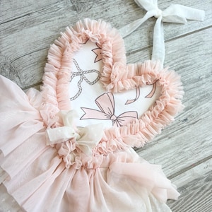 May include: A pink tulle skirt with a heart-shaped fabric applique. The applique has a white background with pink bows and a white ribbon.