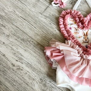 以下が含まれることがあります： ピンクと白のベビー服で、ハート型の胸元に花柄とウサギのプリントが施されています。胸元はフリルになっており、スカートはチュール素材でレースのトリミングが施されています。