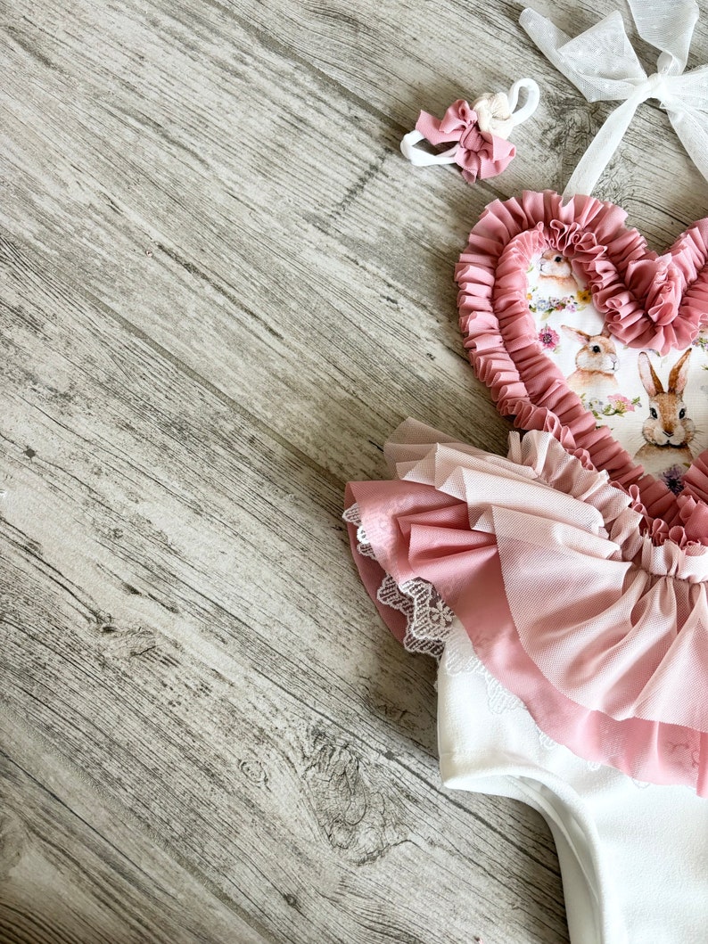 以下が含まれることがあります： ピンクと白のベビー服で、ハート型の胸元に花柄とウサギのプリントが施されています。胸元はフリルになっており、スカートはチュール素材でレースのトリミングが施されています。