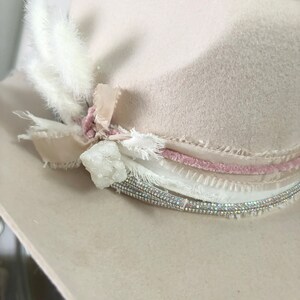 May include: A cream-colored felt hat with a pink velvet band and a white feather accent. The hat is decorated with a crystal and white lace.