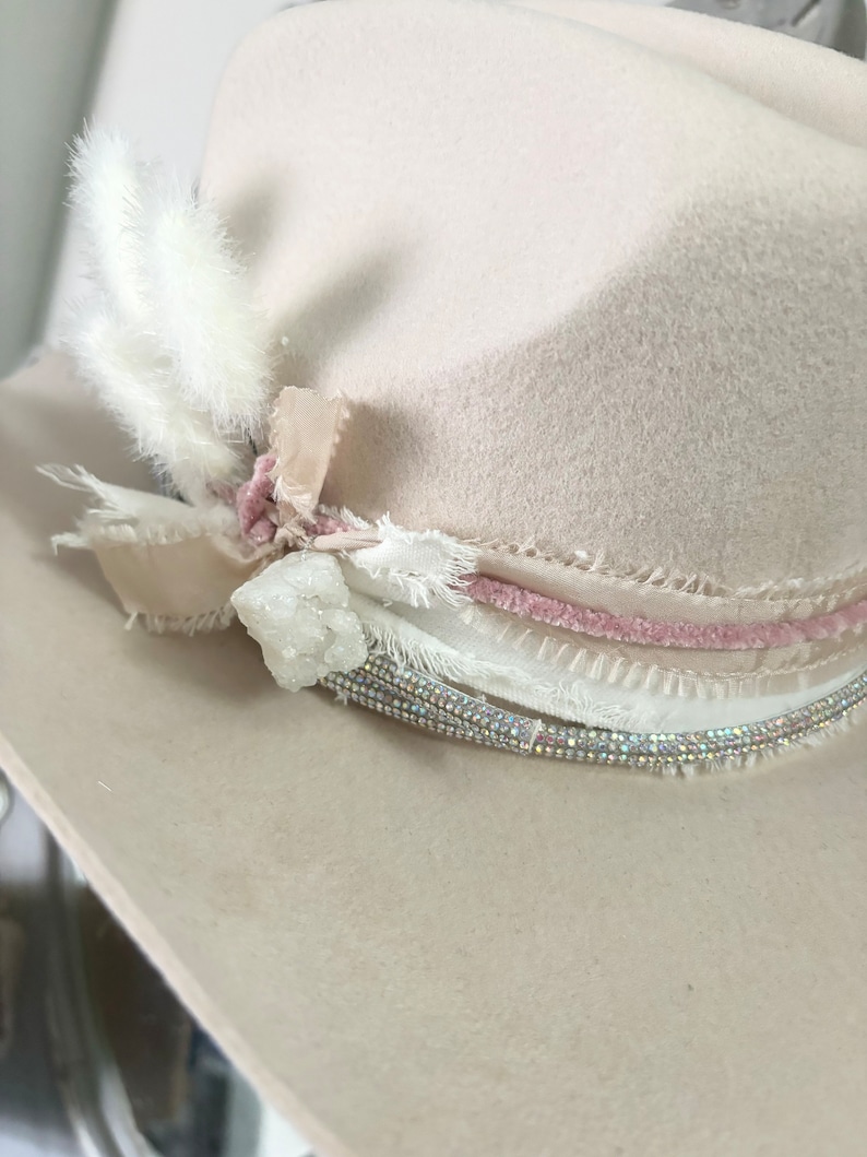 May include: A cream-colored felt hat with a pink velvet band and a white feather accent. The hat is decorated with a crystal and white lace.