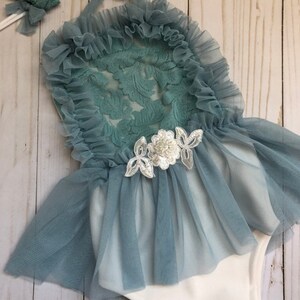 May include: A baby girl's outfit with a teal lace bodice and a tulle skirt. The outfit has a white bodysuit underneath and a matching headband with a pearl embellishment.