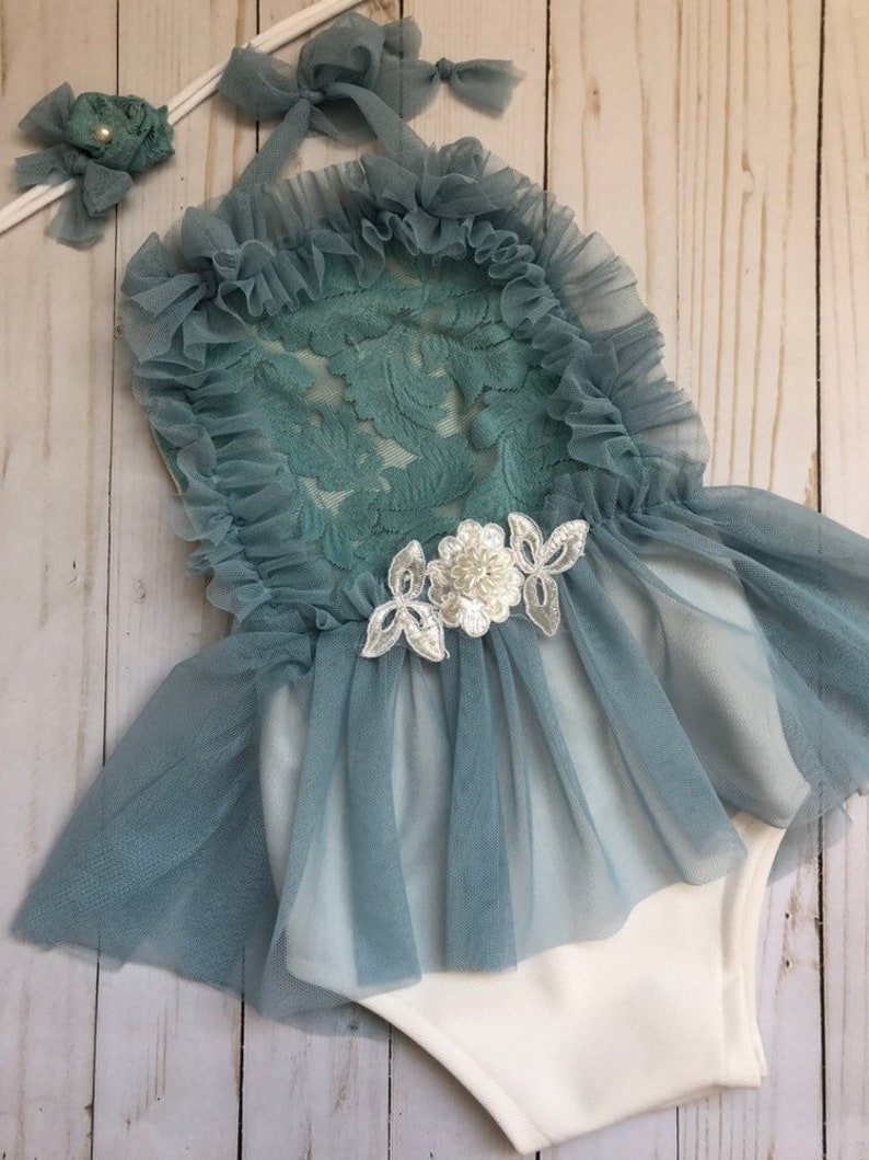 May include: A baby girl's outfit with a teal lace bodice and a tulle skirt. The outfit has a white bodysuit underneath and a matching headband with a pearl embellishment.