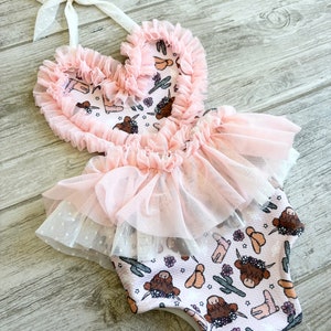 May include: Pink baby onesie with a heart-shaped bodice and a tulle skirt. The onesie has a print of brown highland cows, cacti, and cowboy boots.