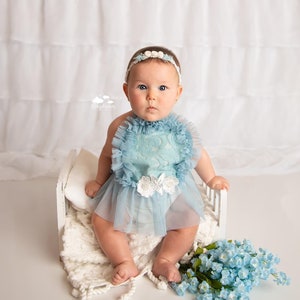 May include: A baby girl wearing a light blue tulle dress with lace details sits on a white bed with a white blanket. She is wearing a white headband with pearl accents. A bouquet of blue flowers is on the floor next to the bed.