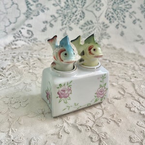 Puede incluir: Un par de saleros y pimenteros de cerámica con forma de pez con detalles florales. Los saleros son blancos con rosas rosas y hojas verdes. Los peces son azules, verdes y amarillos con ojos negros.