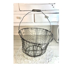 May include: A large, vintage wire basket with a wooden handle. The basket is made of woven wire and has a round, open shape. It is sitting on a white doily.