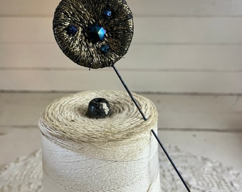 May include: A black and gold round pin with black rhinestones. The pin is attached to a black metal pin back and is sitting on a spool of white thread.