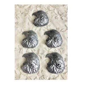 Pode incluir: Cinco pingentes de cabeça de águia prateados com uma superfície texturizada. Os pingentes têm um diâmetro de aproximadamente 2,5 cm.