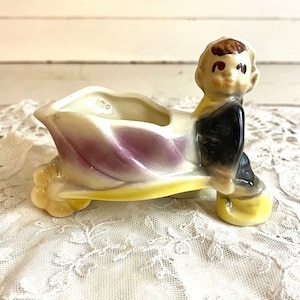 Könnte beinhalten: Eine Keramikfigur eines Menschen, der einen Schubkarren-Pflanztopf schiebt. Der Topf ist in den Farben Lila, Gelb und Weiß bemalt. Die Figur trägt ein schwarzes Outfit und hat braunes Haar.