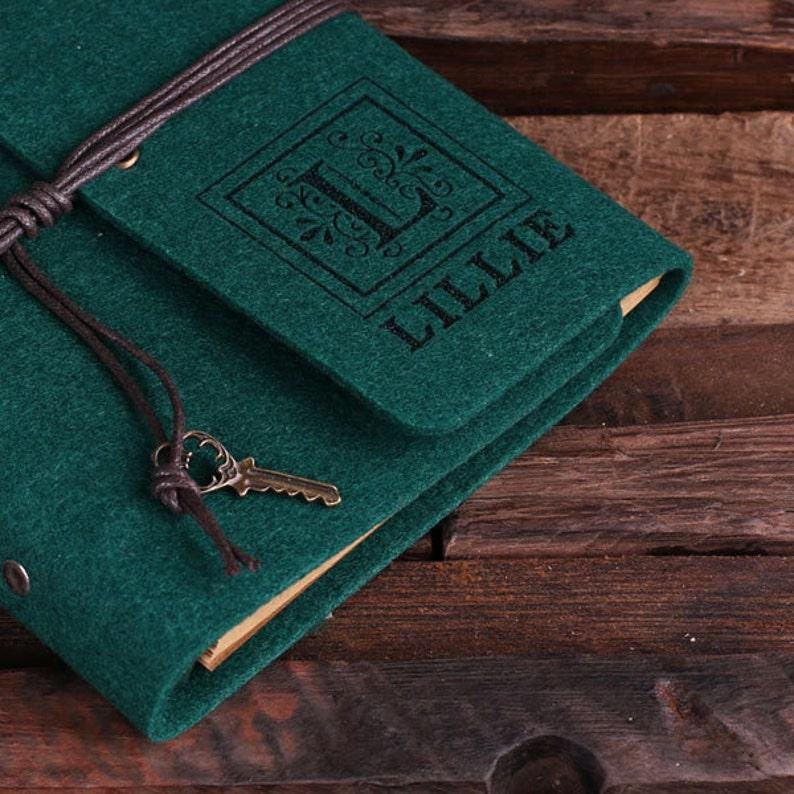 May include: A dark green felt journal with a brown leather strap and a key charm. The journal has a personalized monogram with the letter "L" and the name "Lillie" printed on the front.