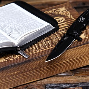 Pu&ograve; includere: Un coltello pieghevole nero con la scritta "Ollie" sul manico &egrave; appoggiato su una superficie di legno accanto a un libro aperto in una custodia nera. Le pagine del libro sono visibili e la custodia ha un tirazip a forma di croce.