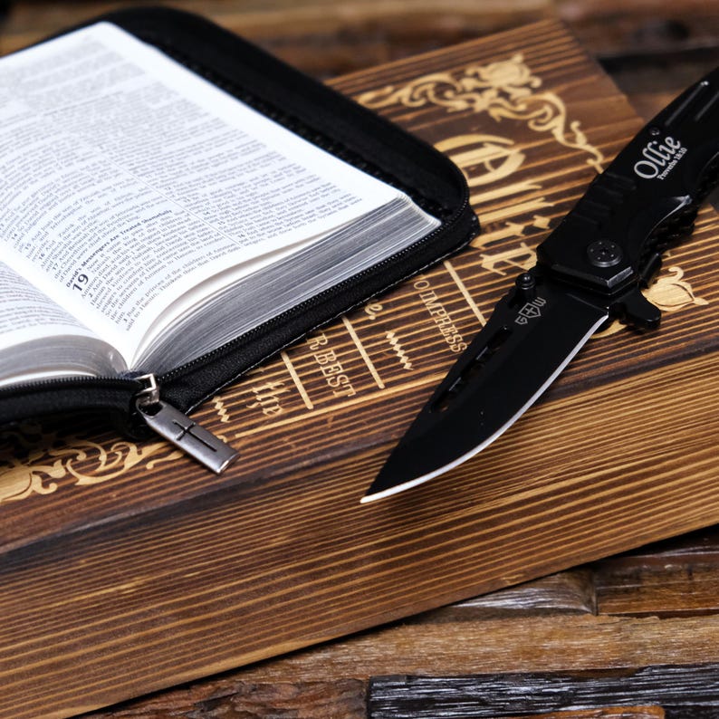 Pu&ograve; includere: Un coltello pieghevole nero con la scritta "Ollie" sul manico &egrave; appoggiato su una superficie di legno accanto a un libro aperto in una custodia nera. Le pagine del libro sono visibili e la custodia ha un tirazip a forma di croce.