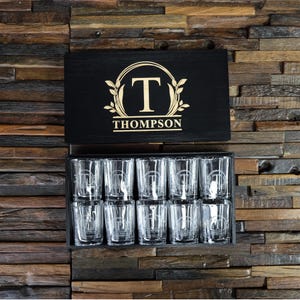 May include: A black wooden box with a gold monogrammed lid that says "T THOMPSON" and contains 12 clear shot glasses with a gold monogram.
