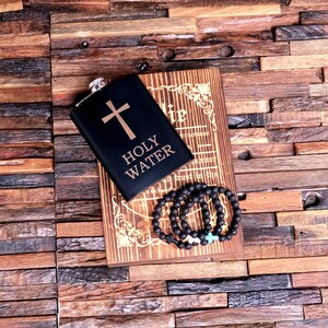 May include: Black flask with a gold cross and the words "Holy Water" engraved on it. The flask is sitting on a wooden box with a book inside. A black beaded bracelet with a cross charm is on top of the box.