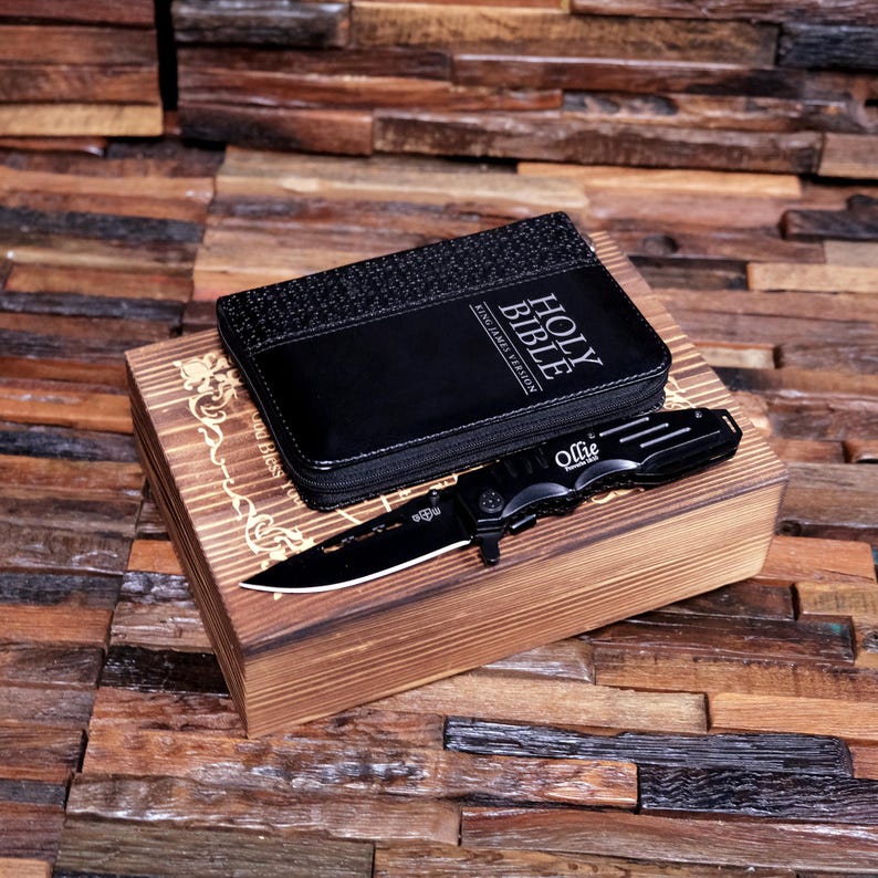Pu&ograve; includere: Un coltello pieghevole nero con la scritta "Ollie" sul manico poggia su una scatola di legno. Una custodia nera con cerniera con "HOLY BIBLE KING JAMES VERSION" &egrave; sopra la scatola. La scatola ha un design decorativo.