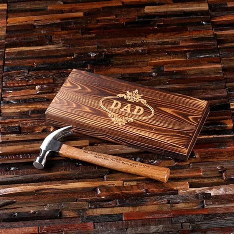 Pu&ograve; includere: Una scatola di legno con la parola "DAD" incisa su di essa. La scatola &egrave; appoggiata su una superficie di legno. Un martello con manico di legno &egrave; accanto alla scatola. Il manico del martello ha il testo "Pap&agrave;, grazie per essere sempre l&igrave; con tutto quello che ti amiamo. I ragazzi. Buon Natale." inciso su di esso.