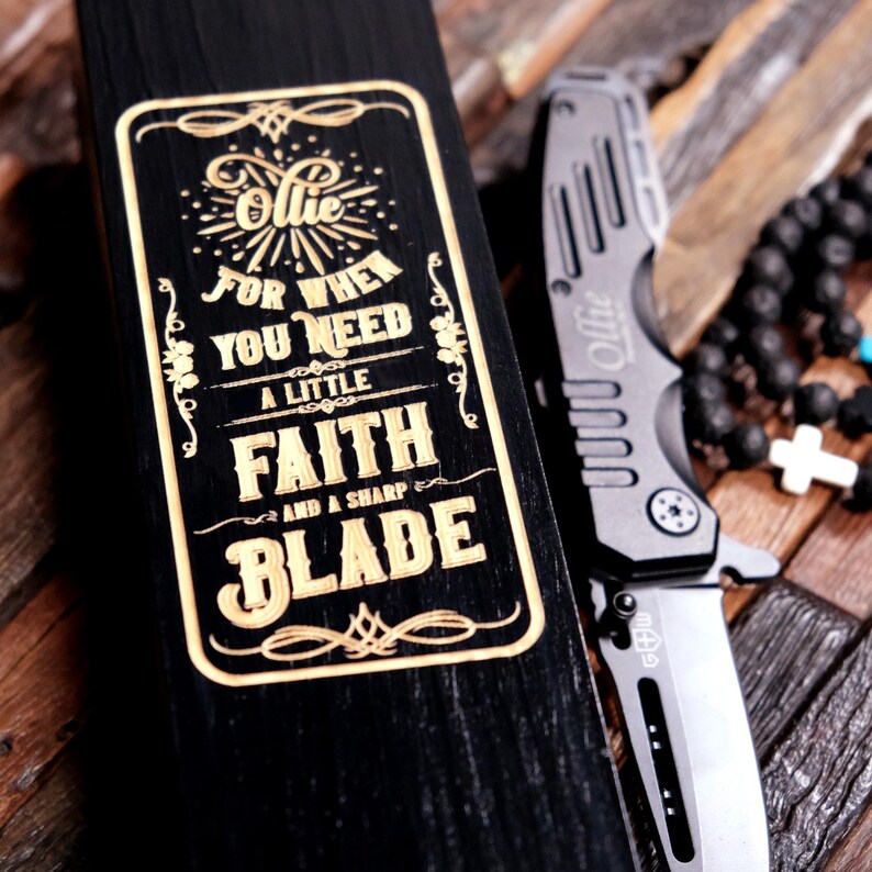 May include: A black wooden box with a gold engraved inscription that reads "Ollie for when you need a little faith and a sharp blade".