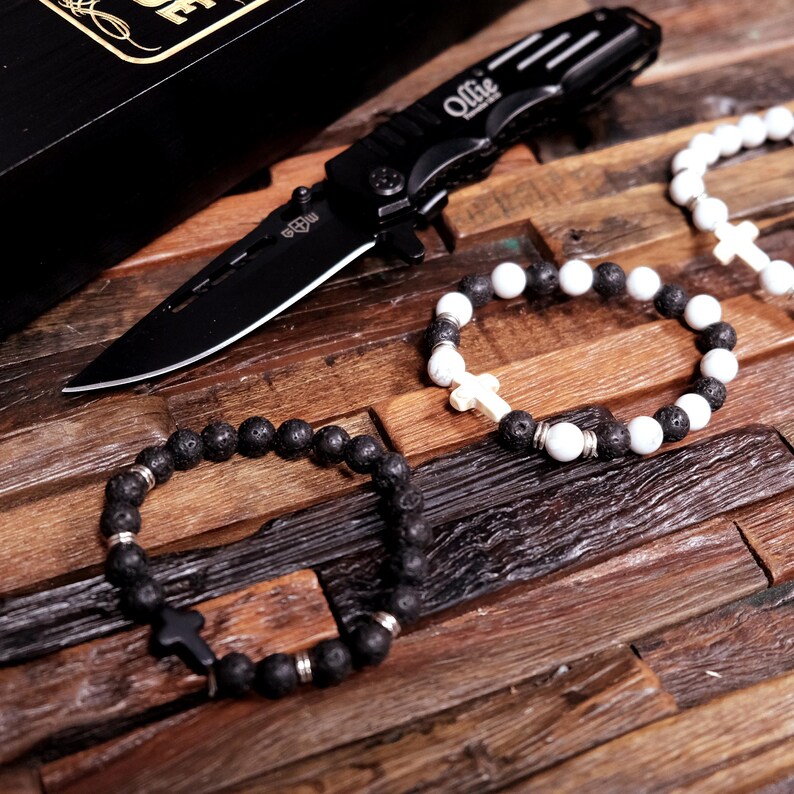 May include: A gift set with a black pocket knife, a black lava rock bracelet with a silver cross charm, and a white and black beaded bracelet with a silver cross charm. The knife has the name "Ollie" engraved on it.