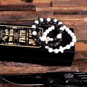 May include: A black box with a gold inscription that reads "For when you need a little faith and a sharp blade".  Three black and white beaded bracelets with cross charms are displayed on top of the box. A black pocket knife with a silver blade is lying on a wooden surface.