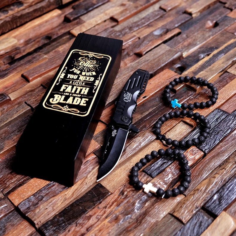 May include: A black folding knife with a black box that says "Ollie For When You Need A Little Faith And A Sharp Blade". Three black beaded bracelets with silver cross charms.