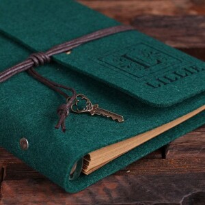 May include: A dark green felt journal with a brown leather strap and a key charm. The journal has a personalized monogram with the letter "I" and the name "Lillie" embossed on the front cover.